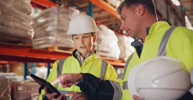 Dois profissionais com equipamentos de segurança analisando dados em um tablet dentro de um armazém logístico, representando a emissão de certificados para exportação e o COD Brasil.